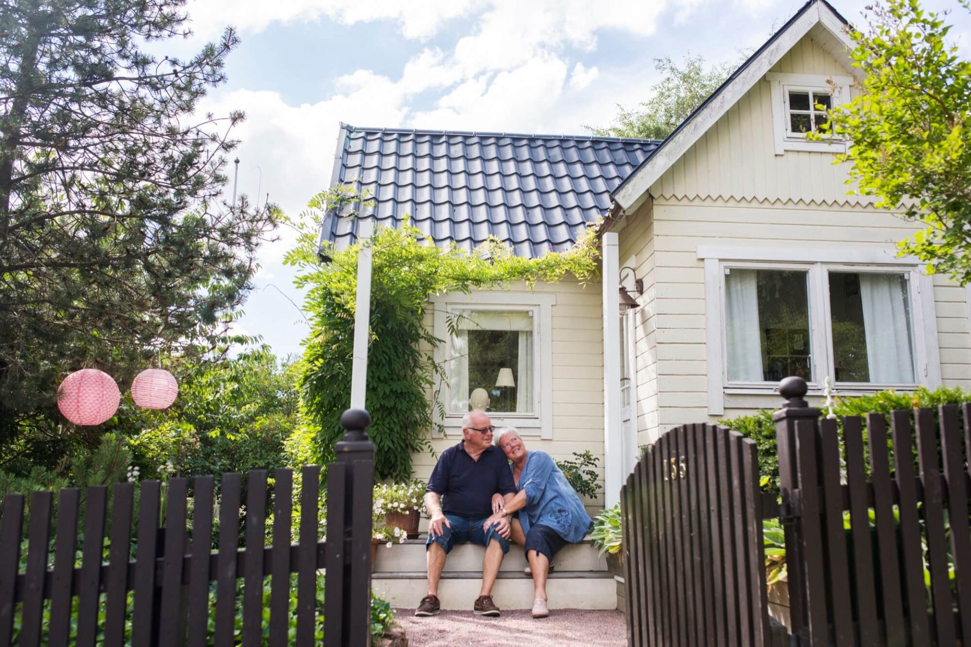 En bild av lugn, ett ljusgult trähus med svart tak, omfamnat av naturens grönska, där ett äldre par tar det lugnt på verandan